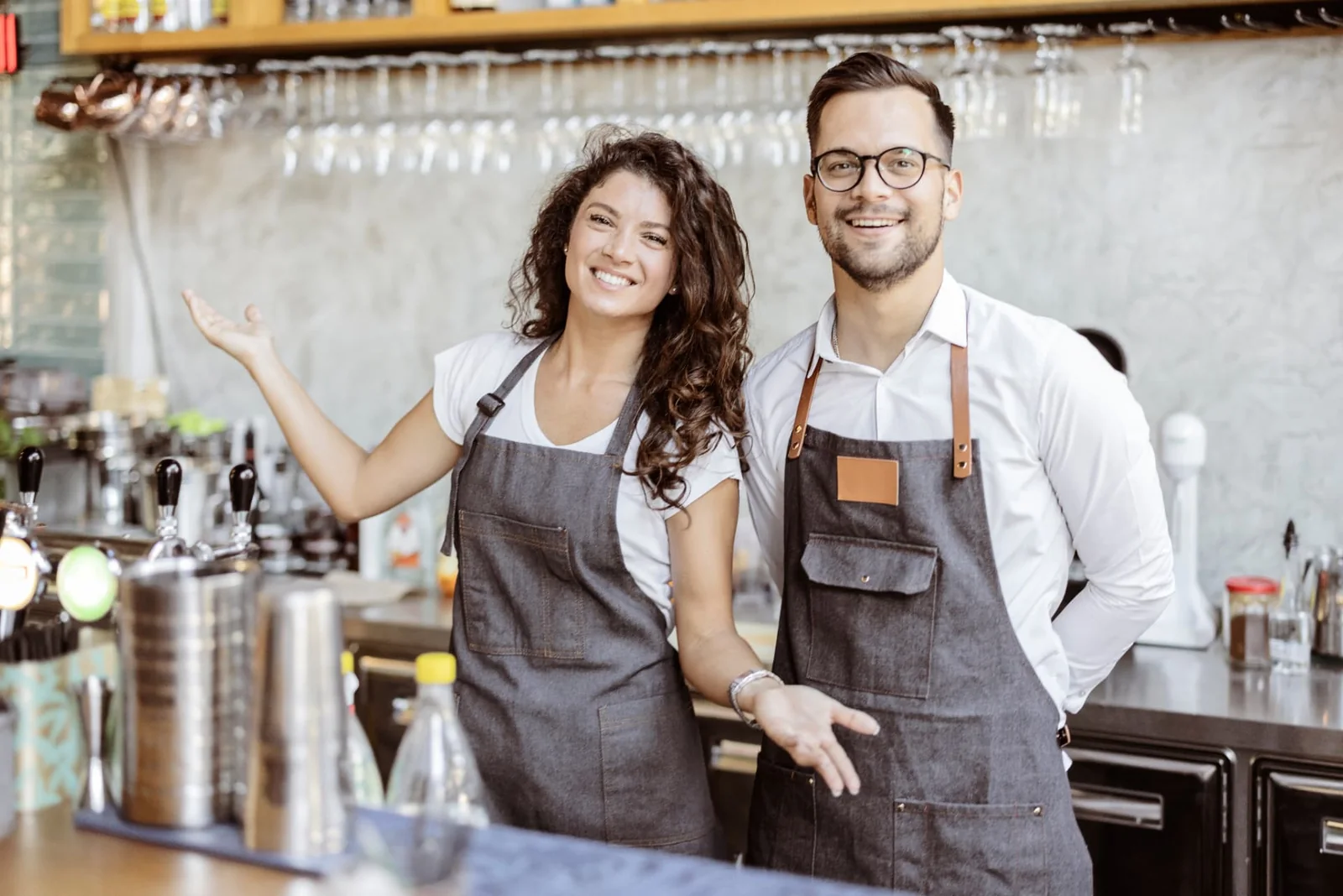 Two smiling café owners in aprons welcoming guests from behind their counter